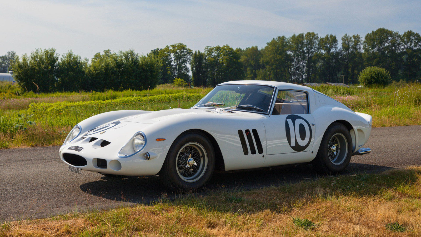 A factory white Ferrari 250 GTO with the number 10 on the door is parked on a rural road, surrounded by grass and trees under a clear sky—a stunning sight reminiscent of a Mecum Kissimmee Auction classic.
