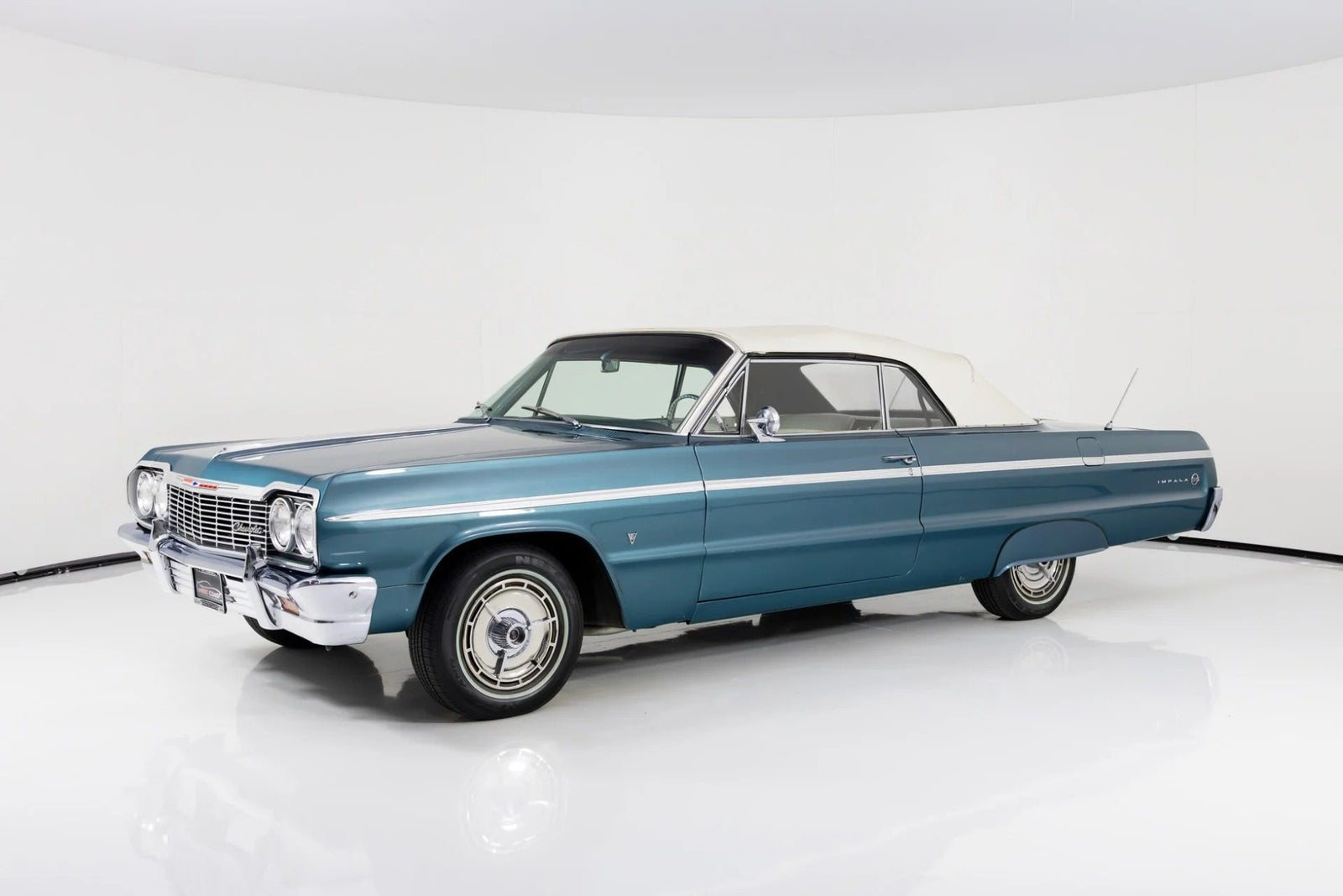 A blue 1964 Chevrolet Impala convertible with a white soft top is parked in a white studio setting, capturing the timeless appeal of classic muscle cars.