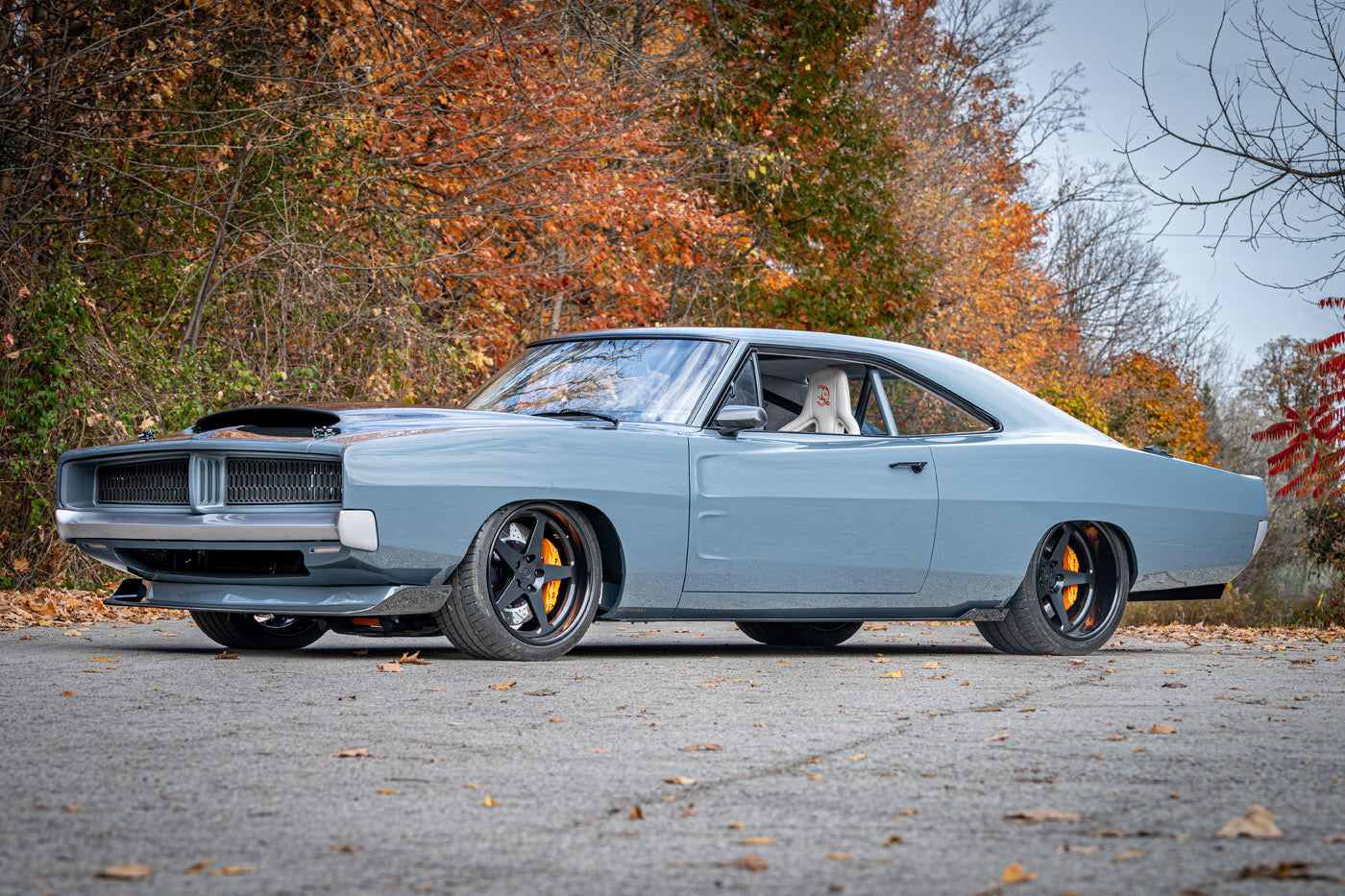 A light blue 1969 Charger classic muscle car with black wheels, yellow brake calipers, and a hood scoop is parked on a paved road with autumn foliage in the background—a true Hellephant-powered icon.