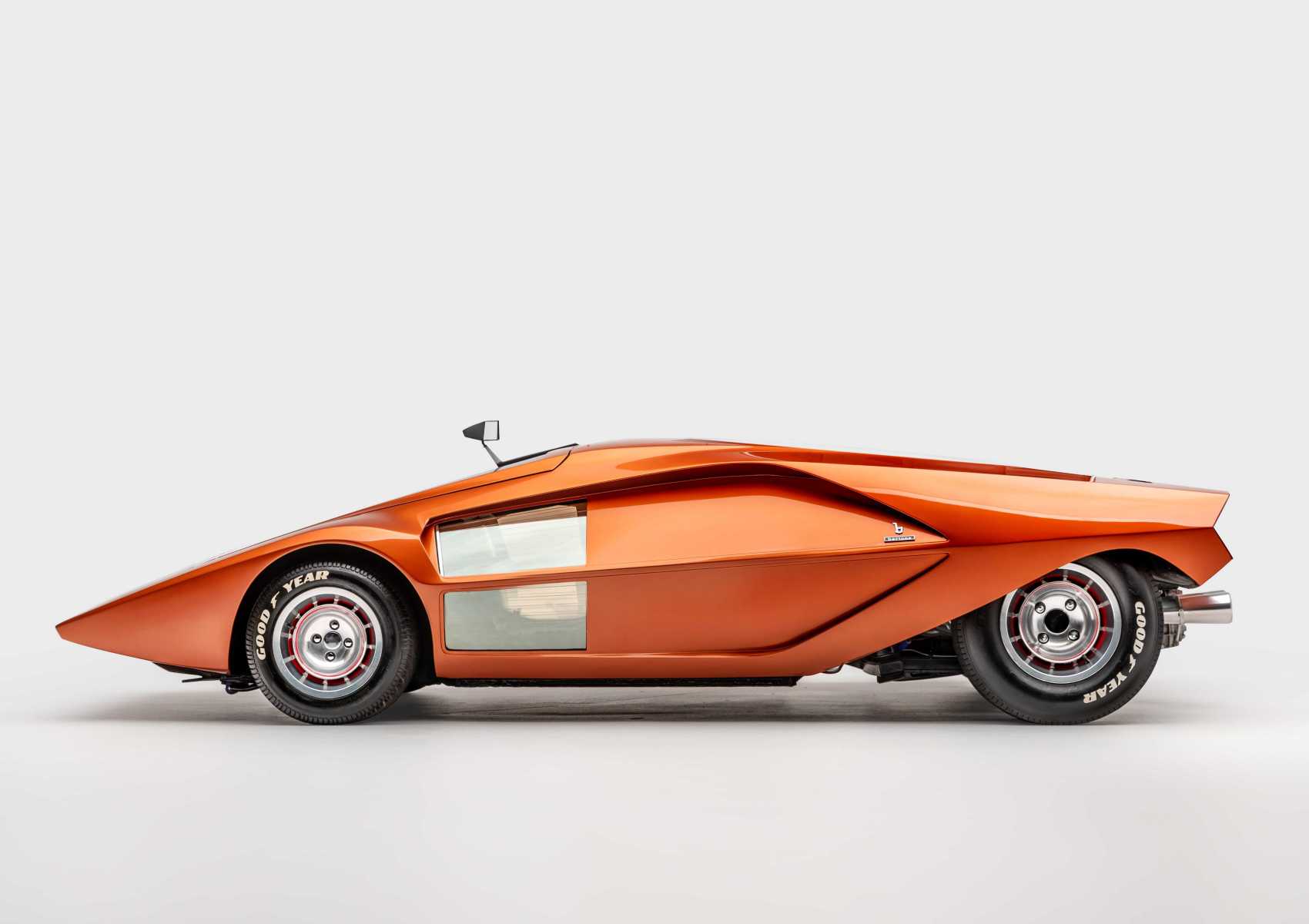 Side view of an orange wedge-shaped concept car with a low, angular profile, small side mirror, visible Goodyear tires, and silver panel detailing, as seen in the exhibit at the Petersen Museum against a plain background.