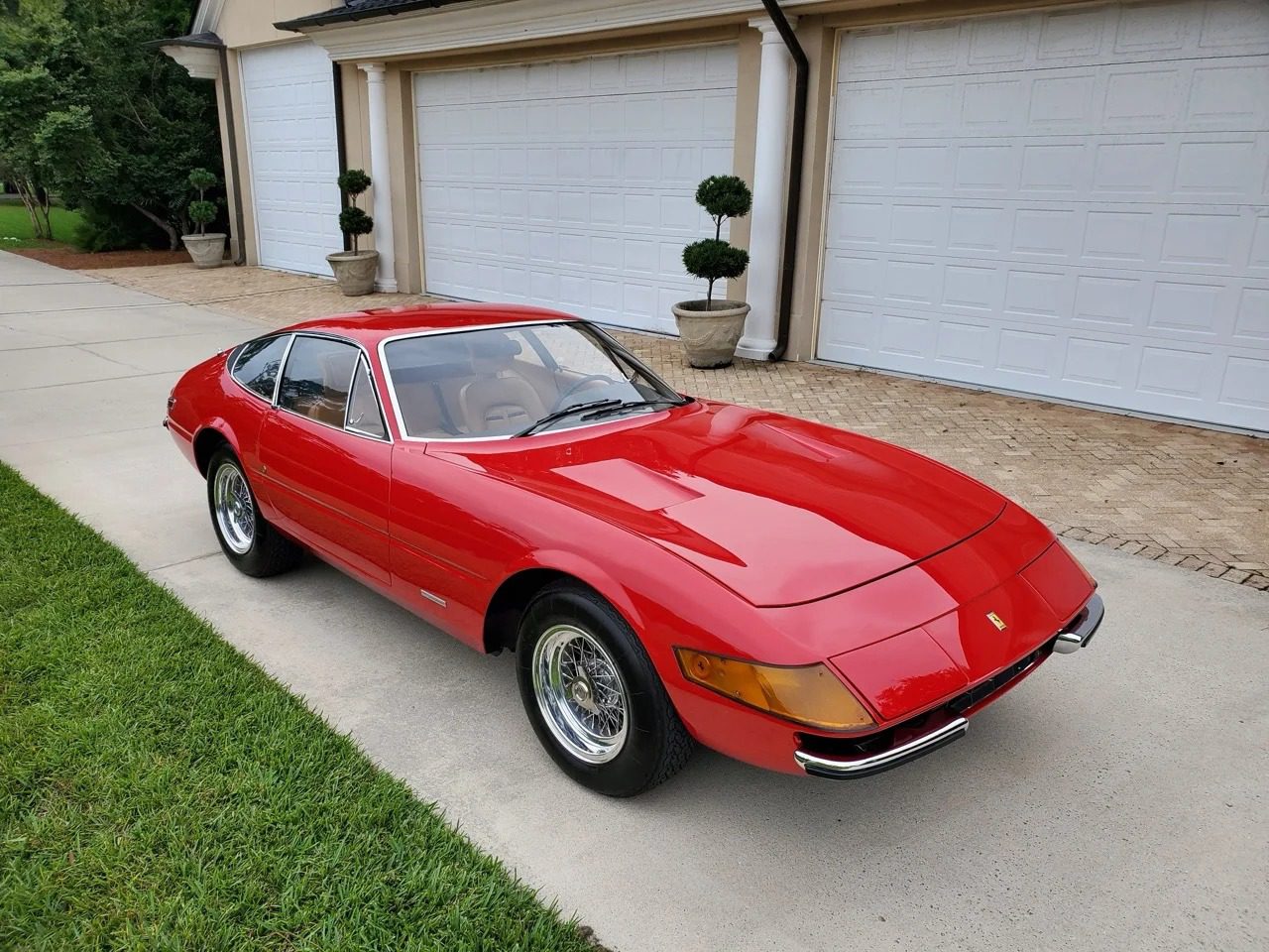 A stunning red vintage sports car, possibly a 1972 Ferrari 365 from the Ferrari Classiche collection, sits gracefully in a driveway. Its beige interior gleams under the sun, with a minimalist garden creating an elegant backdrop.