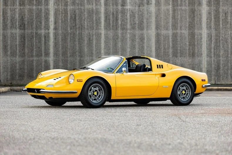 A yellow vintage Ferrari Dino 246 GTS sports car is parked on an empty street in front of a gray concrete wall, showcasing the timeless appeal of classic Ferrari Dino cars.