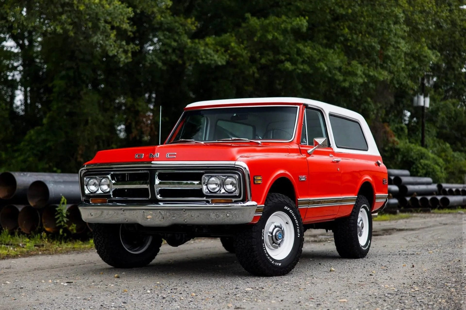 A vintage red and white GMC SUV is parked on a gravel surface with green trees in the background, perfect for classic trucks enthusiasts.