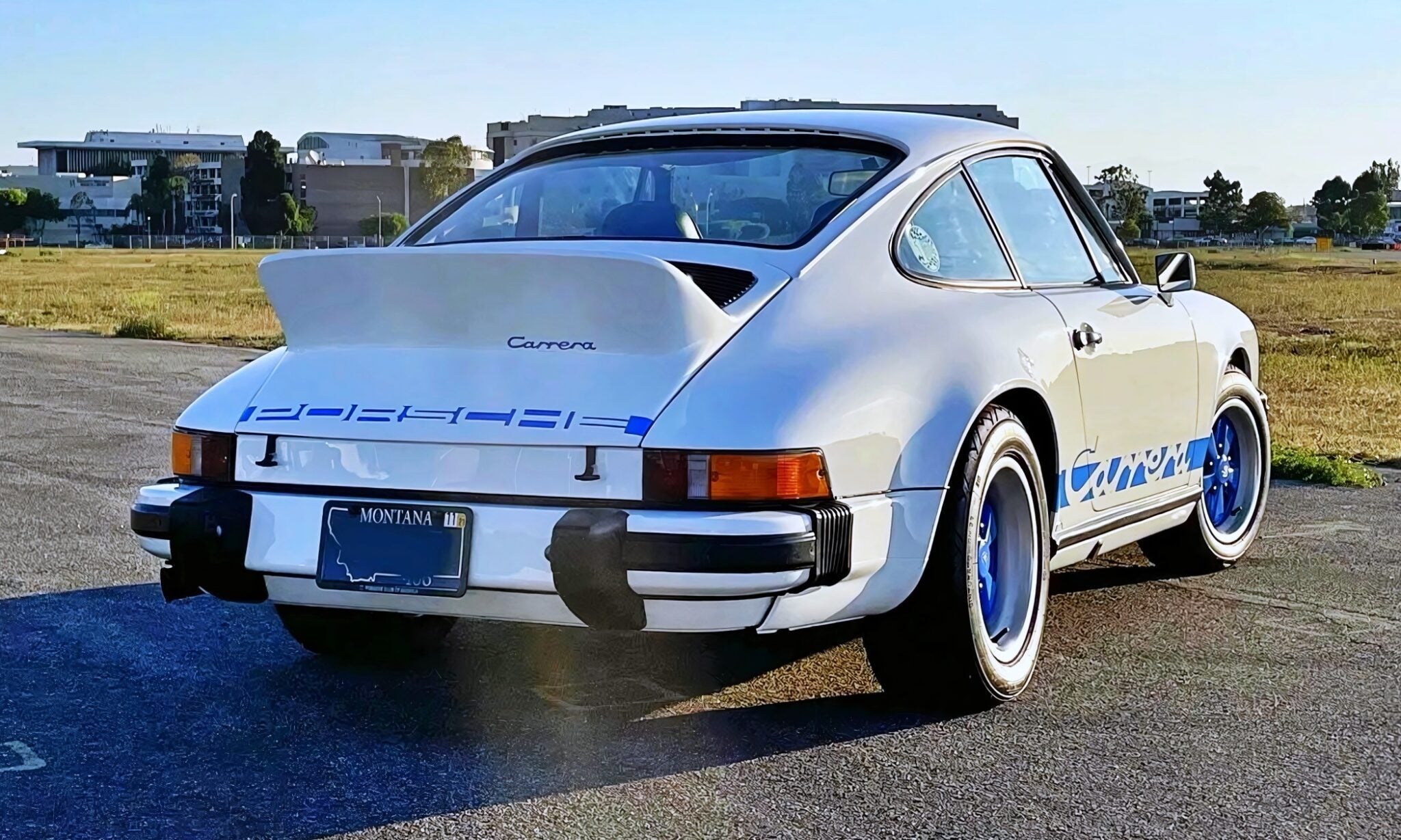 A white classic 1974 Carrera 2.7 MFI for sale, with blue accents and a Montana license plate, is parked on an open lot with grass and buildings in the background—this legend can outrun a Porsche 911 Turbo 3.0.