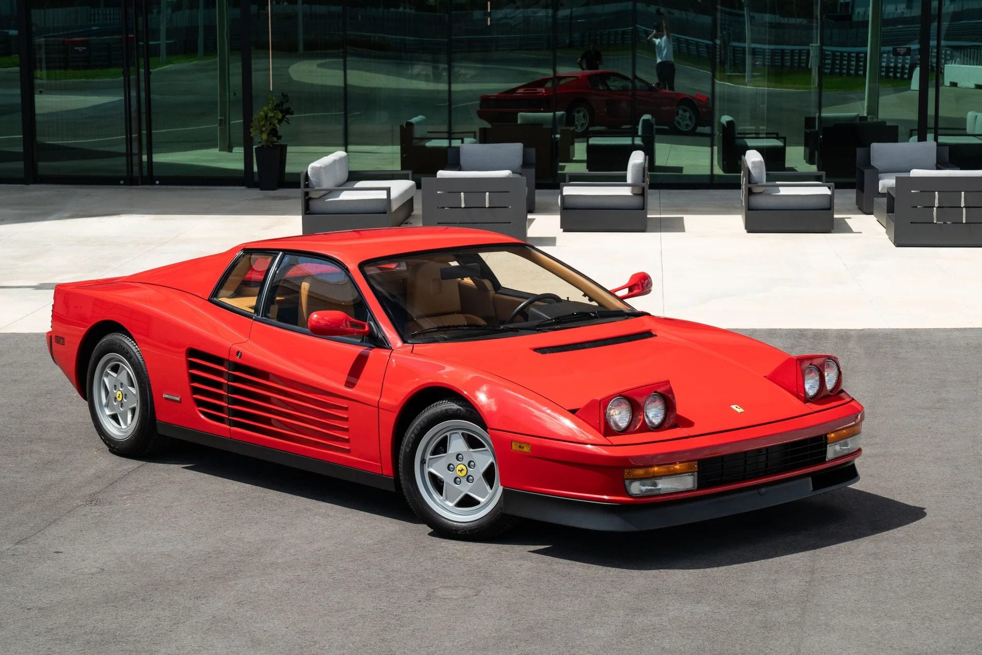 A rare 1990 Ferrari Testarossa sports car is parked on pavement in front of a modern glass building with outdoor seating.