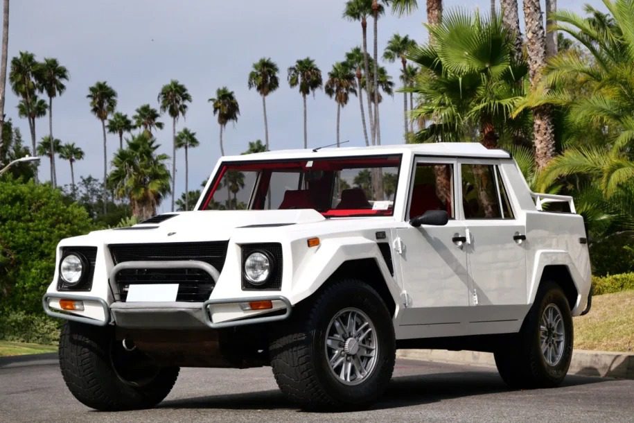 A 1990 Lamborghini LM002, an iconic white off-road vehicle, is parked on a street, surrounded by swaying palm trees and lush greenery. This rare Lamborghini for sale brings adventurous spirit and luxury to any terrain.