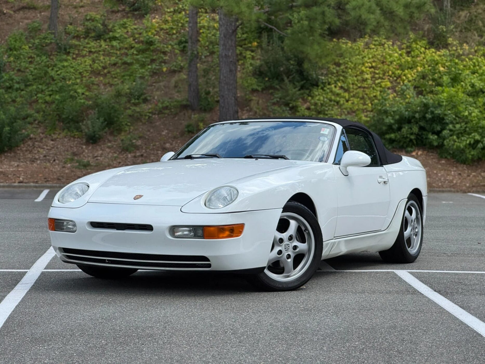 A white Porsche 968 convertible, one of the unsung classics among Transaxle Porsche models, is parked at an angle in an empty outdoor lot with trees and greenery in the background—an ideal car to invest in.