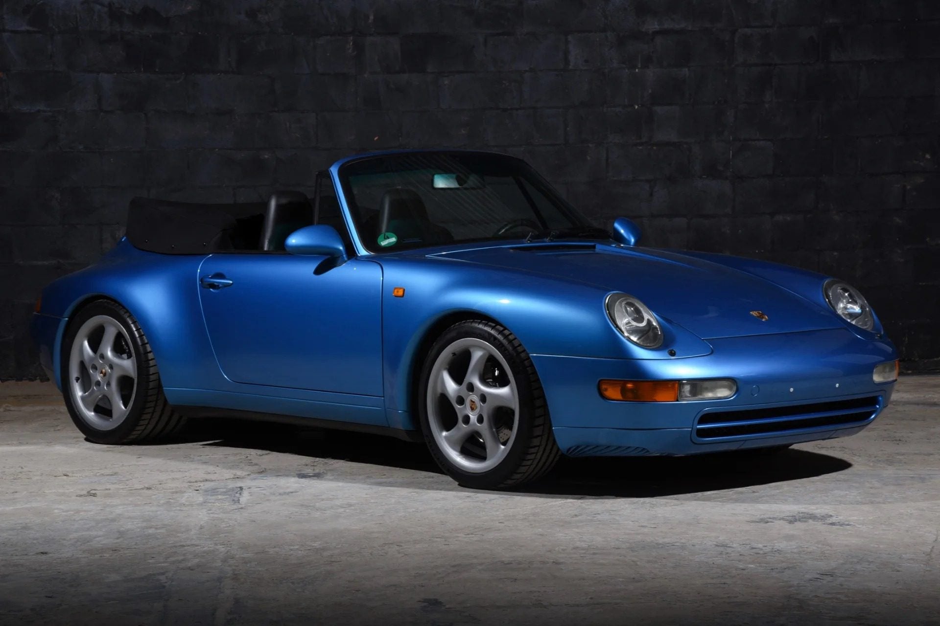 A high-performance blue Porsche 911 convertible is parked indoors on a concrete floor with a dark, textured wall in the background.