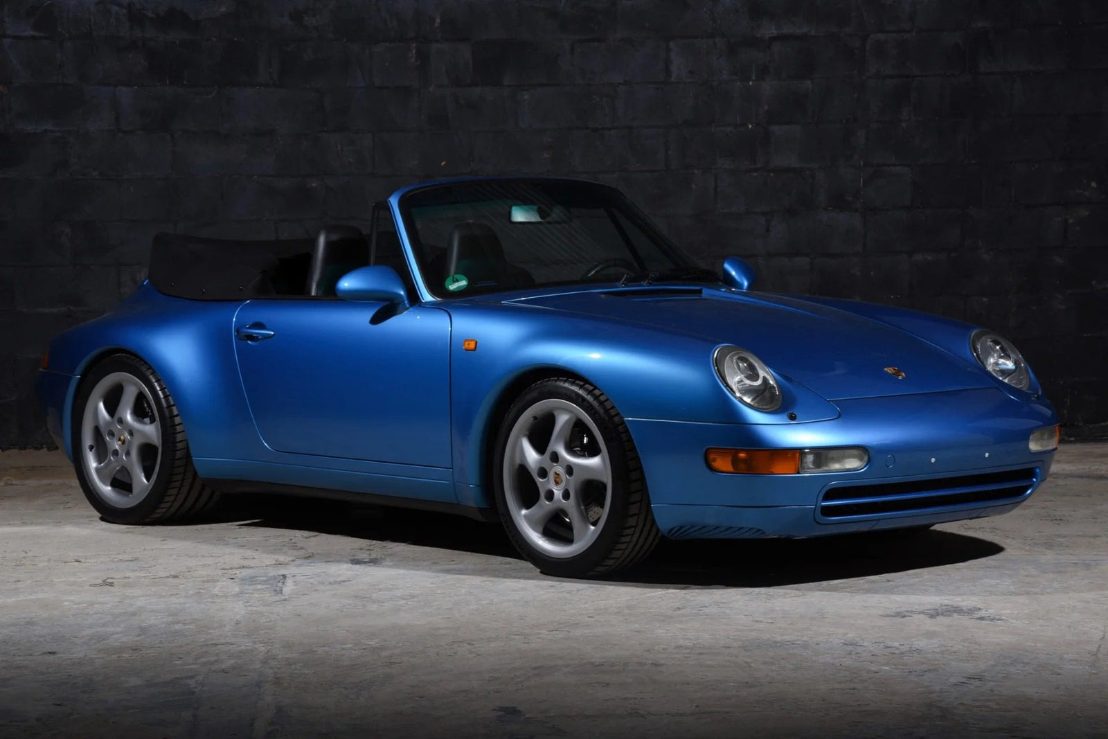 A high-performance blue Porsche 911 convertible is parked indoors on a concrete floor with a dark, textured wall in the background.