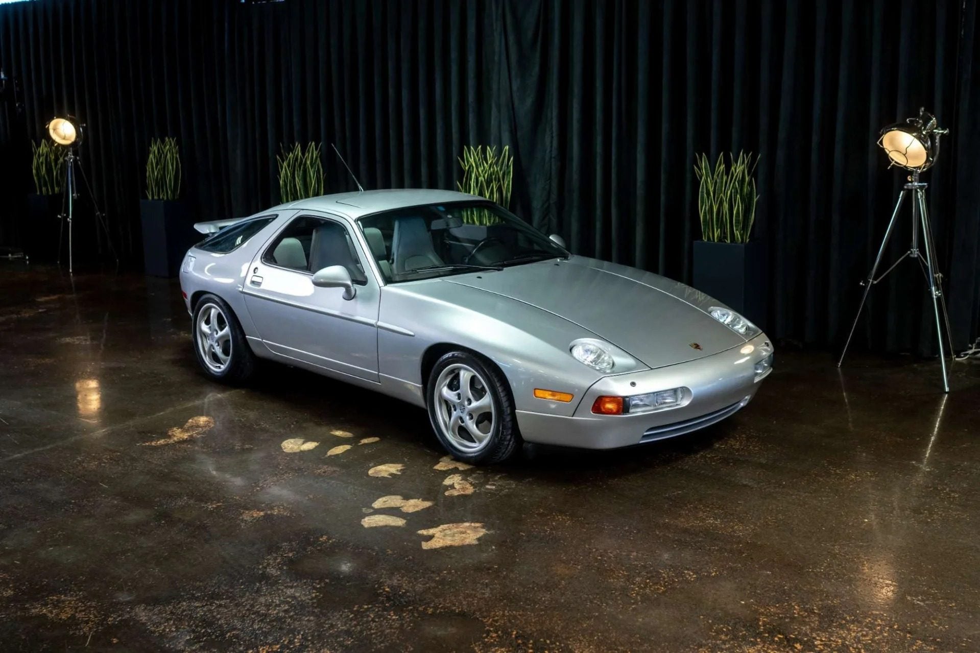 A silver Porsche 928 sports car, a true icon of 90s cars for sale, is parked indoors on a polished floor, with black curtains and potted plants in the background.