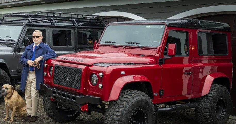 A man in a blue coat and sunglasses stands next to a red off-road vehicle, highlighting the Helderburg Defender Restorations legacy, with a dog sitting nearby. Another similar vehicle, a testament to modern luxury, looms in the background.