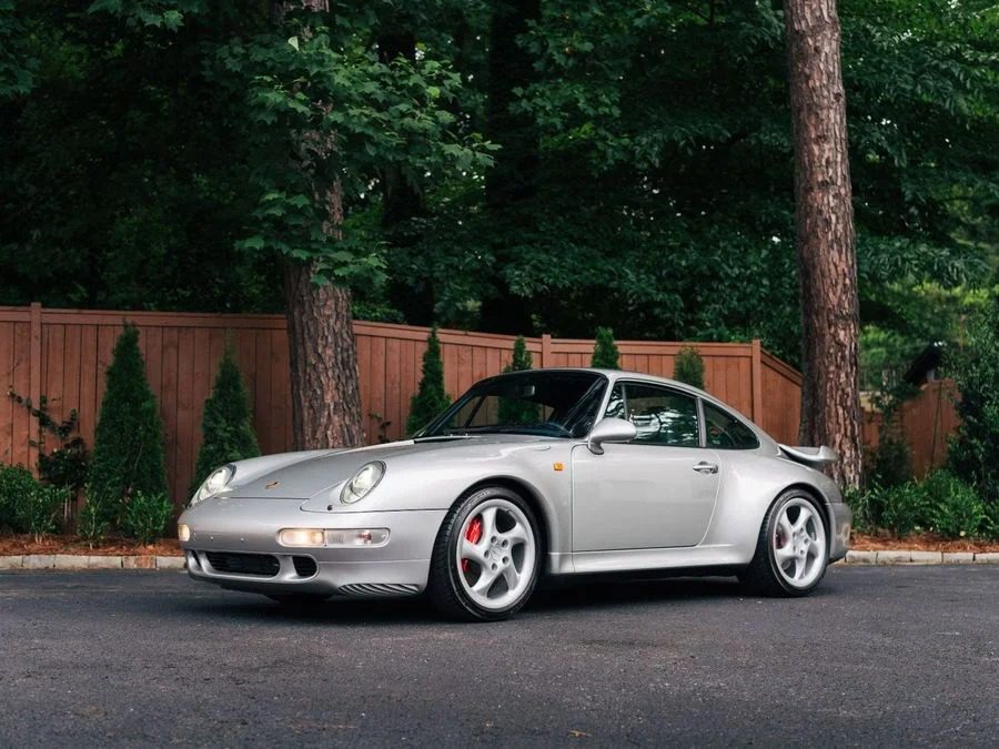 A silver Porsche 911, one of the Best Air-Cooled Porsches, is parked on a paved surface in front of a wooden fence and green trees.