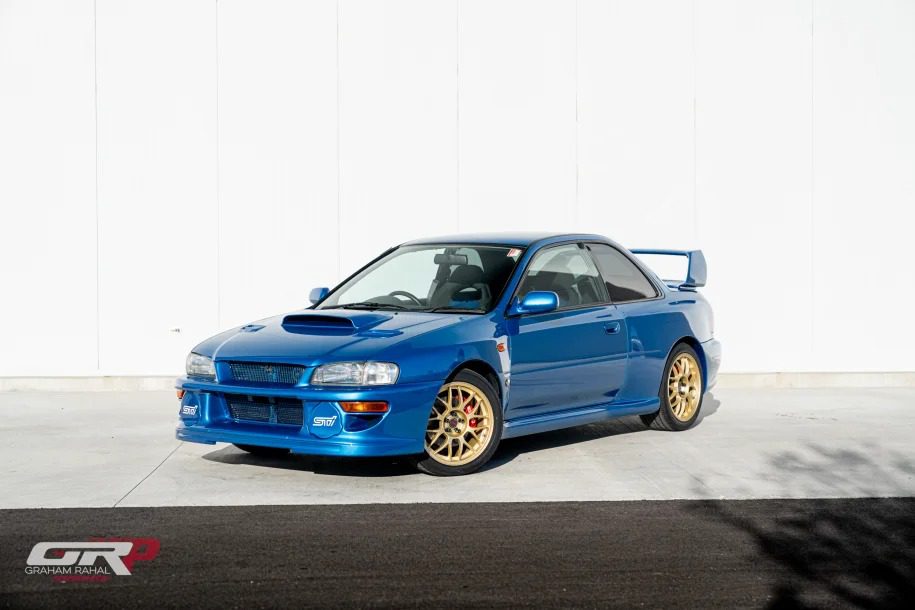 A rare 1998 Subaru Impreza WRX STi 22b with gold wheels is parked in front of a white wall. For sale to collectors and enthusiasts, this blue sports car captures the spirit of a thrilling legacy.