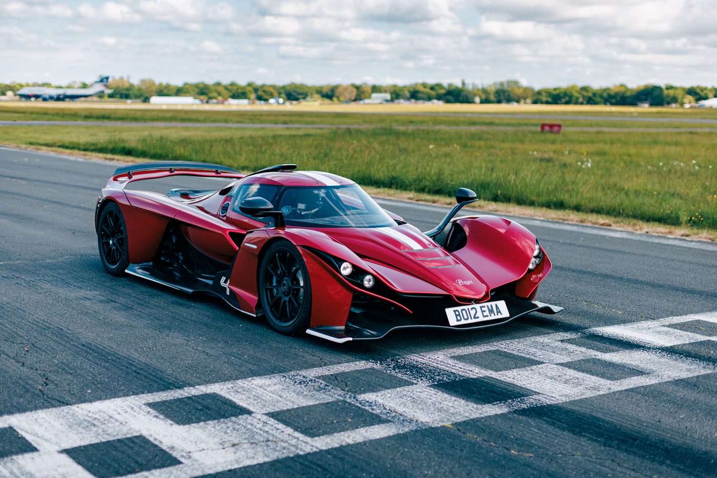 A red supercar is parked on the Top Gear Test Track at the starting line, with grass and trees in the background under a partly cloudy sky, ready to chase a new lap record.