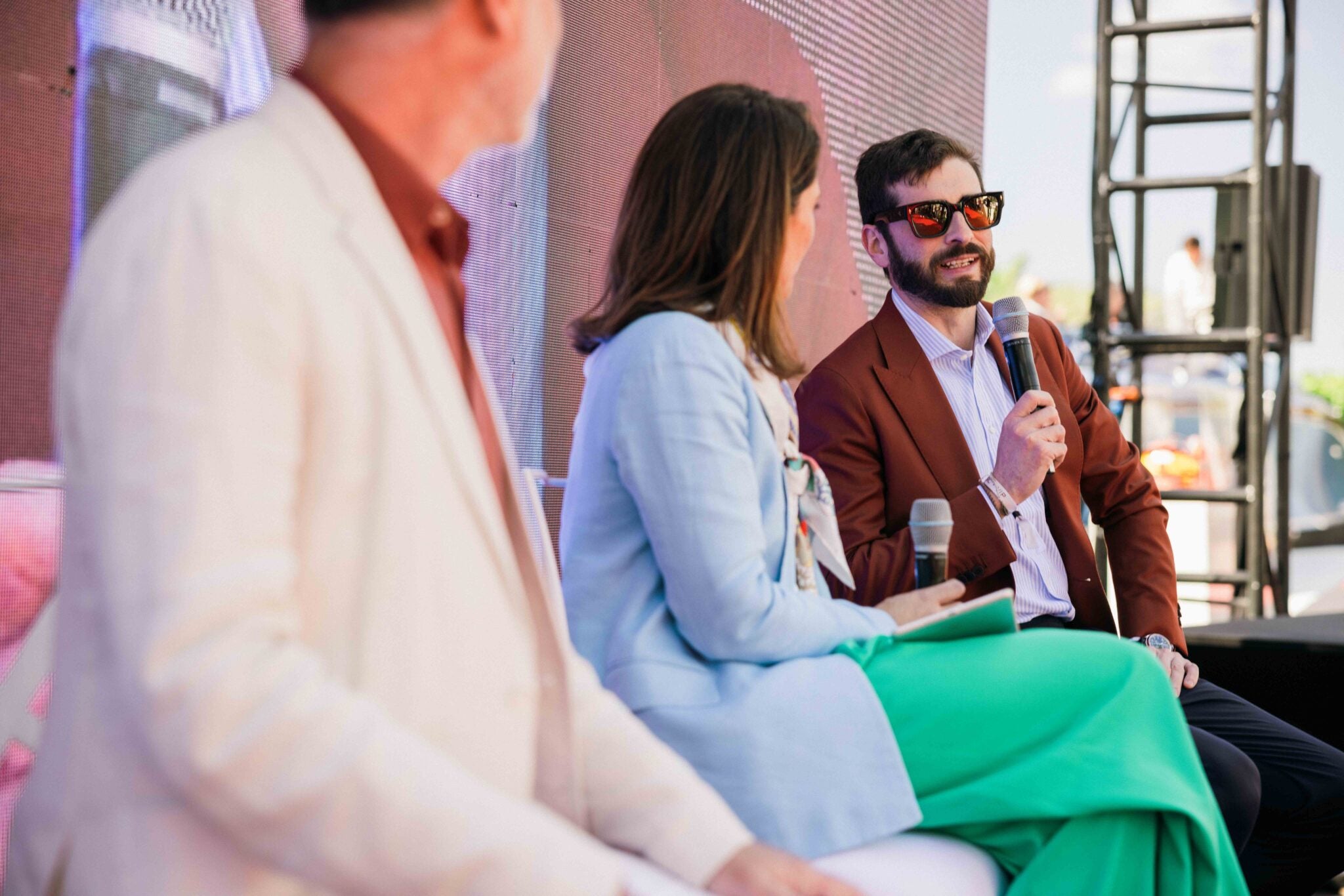 Three people sit on stage during a panel discussion. The man on the right, Enzo Mattioli Ferrari, holds a microphone and speaks, while the other two listen, engaged in the interview about his lasting legacy.