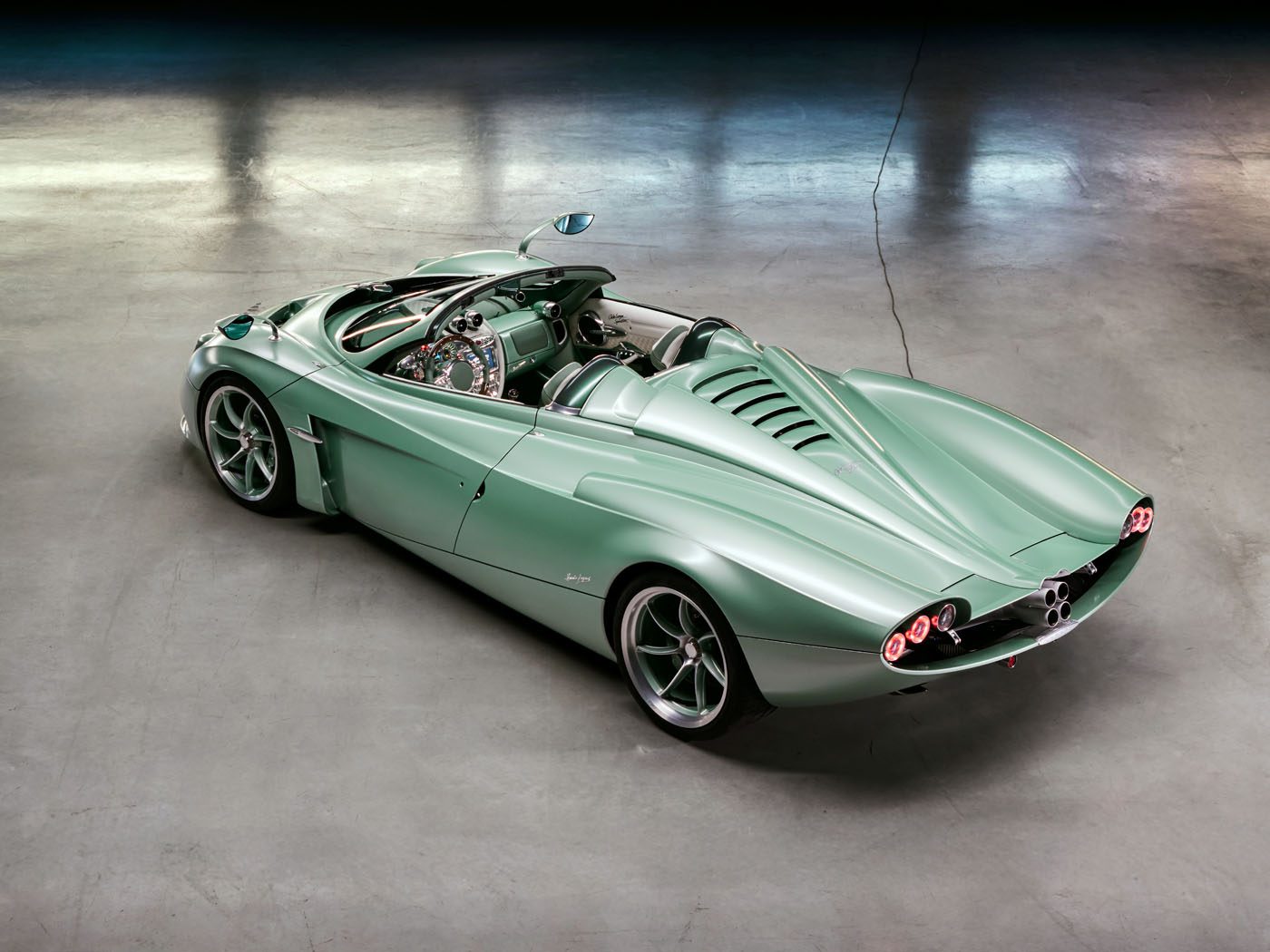 A light green convertible sports car with retro-futuristic design, shown from the rear with an open cockpit and distinctive tail lights, parked on a concrete floor.