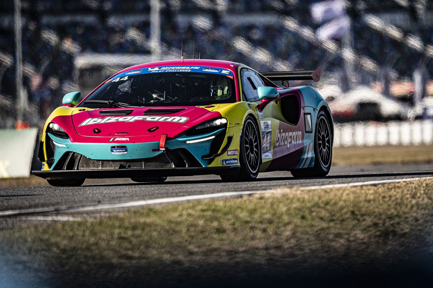 The vibrant McLaren Artura GT4 with "IbizaRam" branding speeds along the track, hinting at its future triumph at the 2025 24 Hours of Daytona. Blurred backgrounds and fencing frame its electrifying presence.