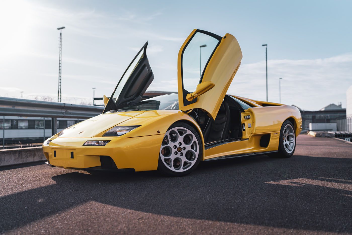 A gleaming yellow Lamborghini Diablo VT 6.0, with its scissor doors dramatically open, rests on a paved surface under the clear blue sky—an icon ready for the spotlight at Broad Arrow Auctions.