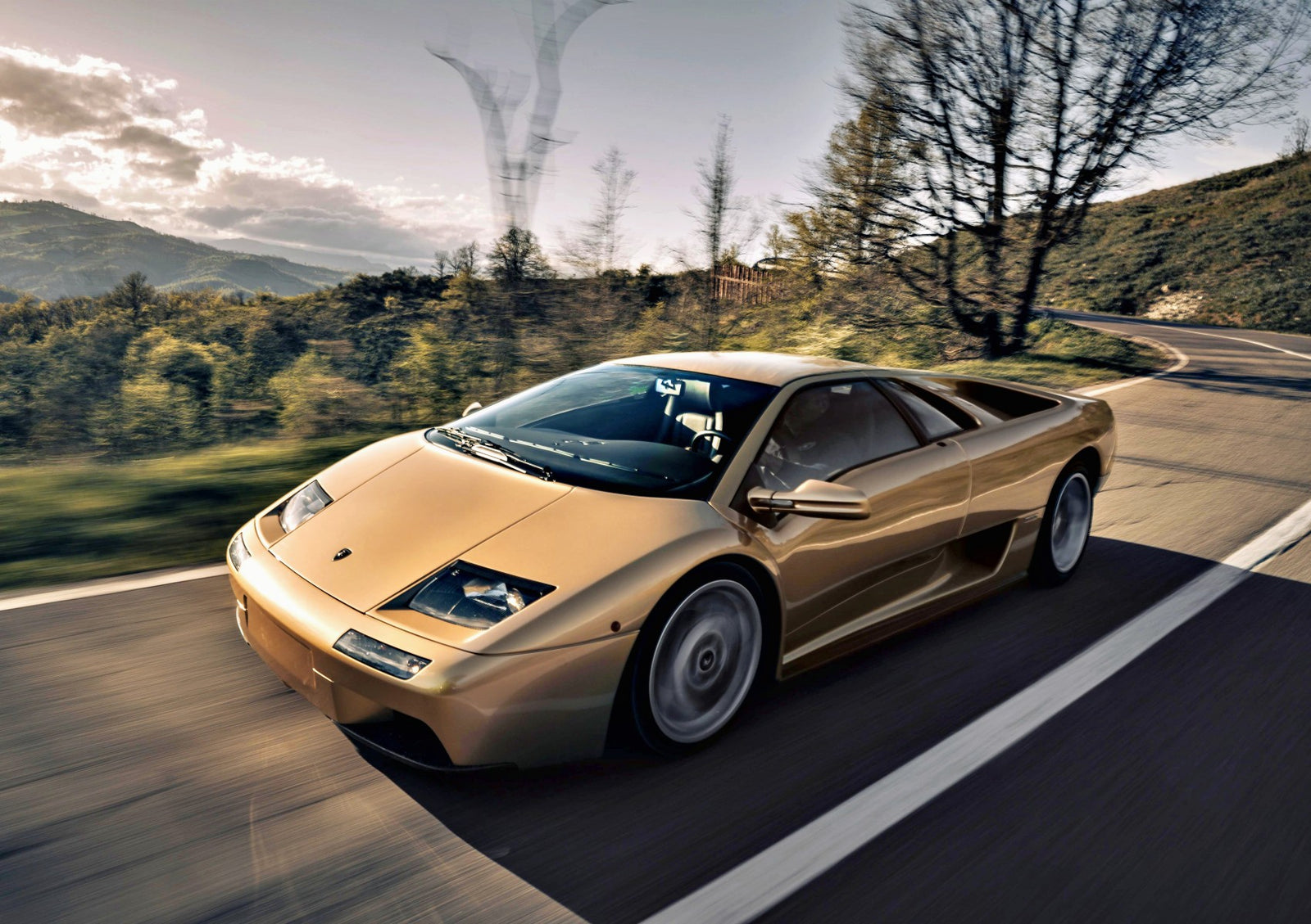 A gold Lamborghini Diablo cruises along a winding Emilia-Romagna road, lined with trees and hills beneath a partly cloudy sky—a perfect scene for fans of vintage Lamborghinis.