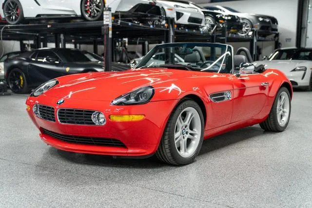 A red convertible sports car, a modern classic, is parked indoors in a sleek garage. It's basking in the spotlight beside other luxury cars on lifts. This open-air beauty could easily be mistaken for a BMW Z8, ready to unleash its charm on the open road.