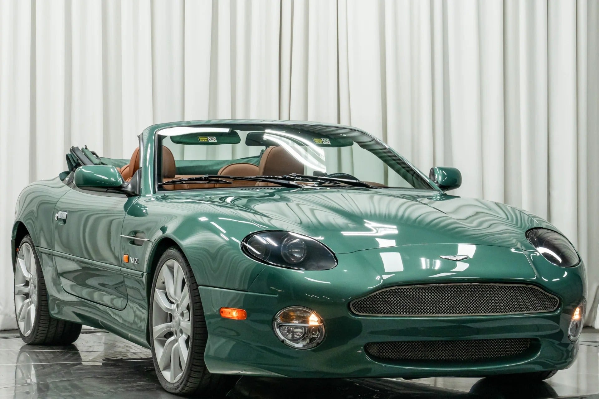A green convertible sports car with the top down is parked indoors in front of white curtains, showcasing one of the classic automotive color combos often sought after by enthusiasts searching for standout cars for sale.