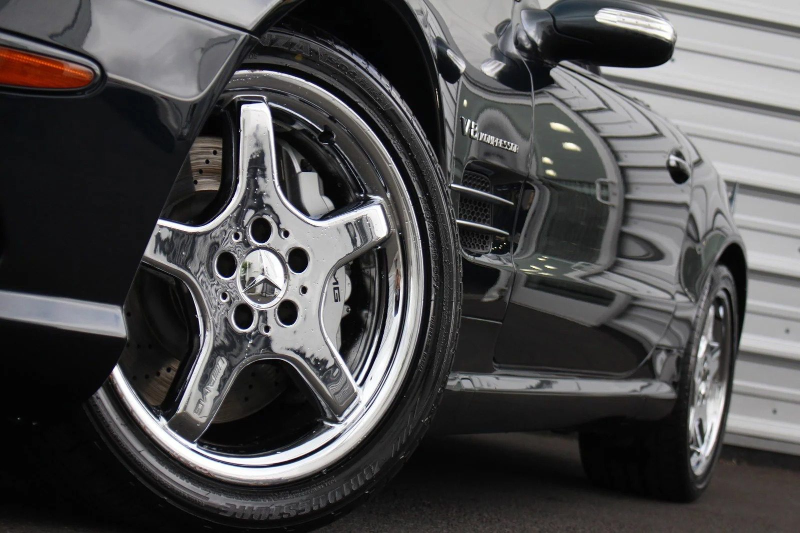 Close-up view of a luxury Mercedes-Benz sports car, focusing on a shiny chrome wheel and part of the front fender, parked near a metal-clad building.