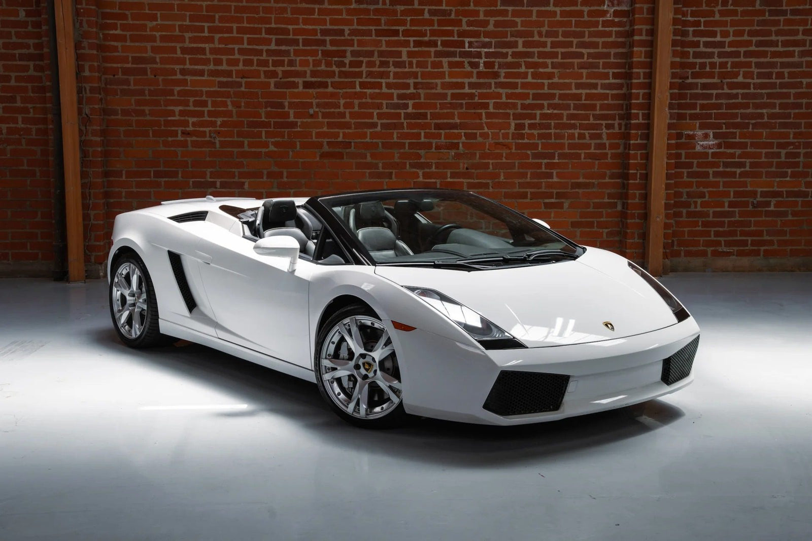 A white Lamborghini Gallardo convertible V10 supercar is parked indoors on a polished floor in front of a brick wall.