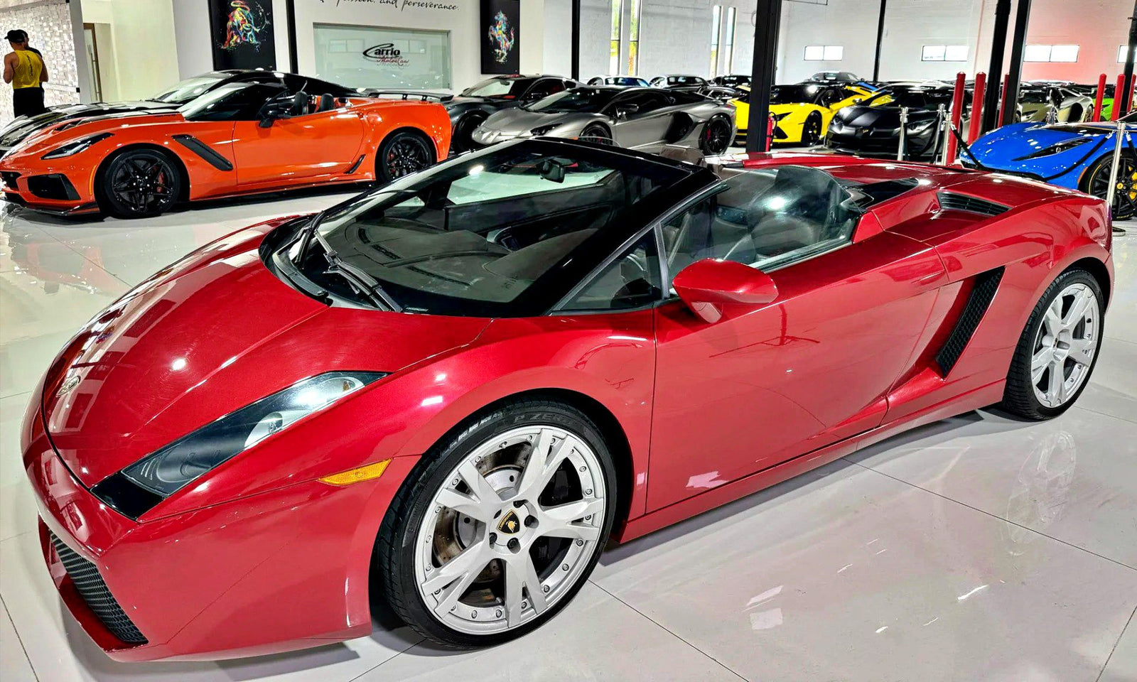 A red Lamborghini Gallardo Spyder convertible is parked indoors among several other luxury sports cars in a showroom, showcasing why it’s considered one of the Best Used Cars For $100000.