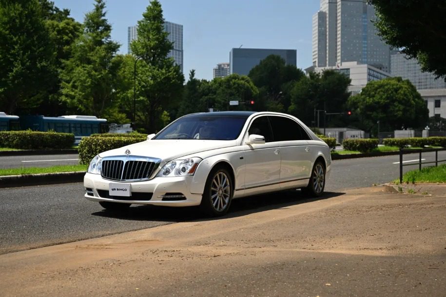 A white sedan, possibly a right-hand drive Maybach 62S Landaulet, is parked on a city street, surrounded by trees and buildings under a clear blue sky.