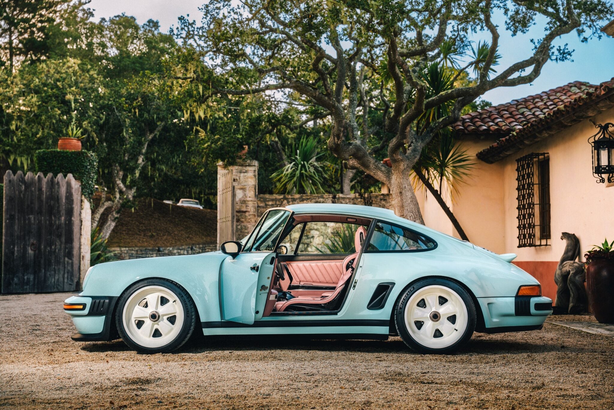 A light blue Reimagined Porsche 911 with white wheels is parked outside a house, the driver’s door open and trees in the background—a classic vision brought to life by Singer Vehicle Design.