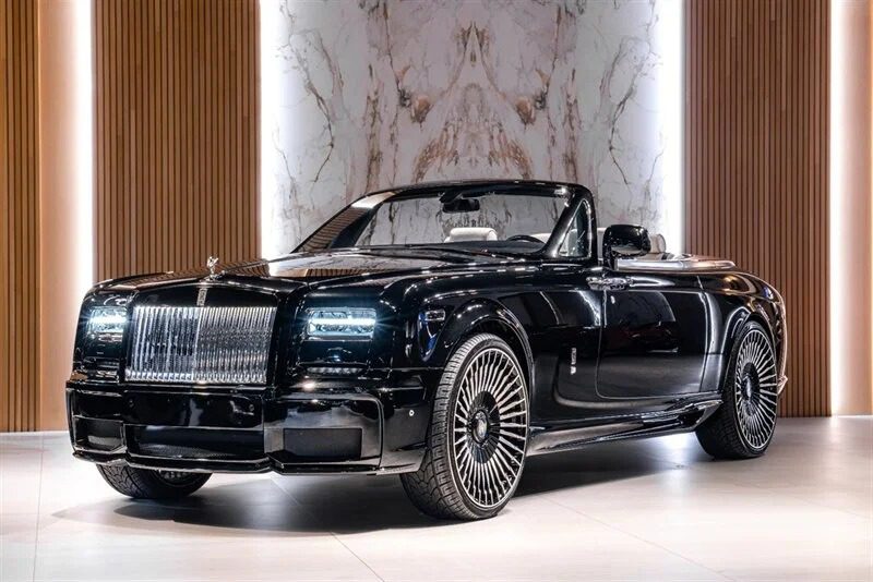 A black Rolls-Royce Phantom convertible, the epitome of a luxury car, is displayed indoors on a light floor with vertical wooden panels and marble walls as the elegant backdrop.