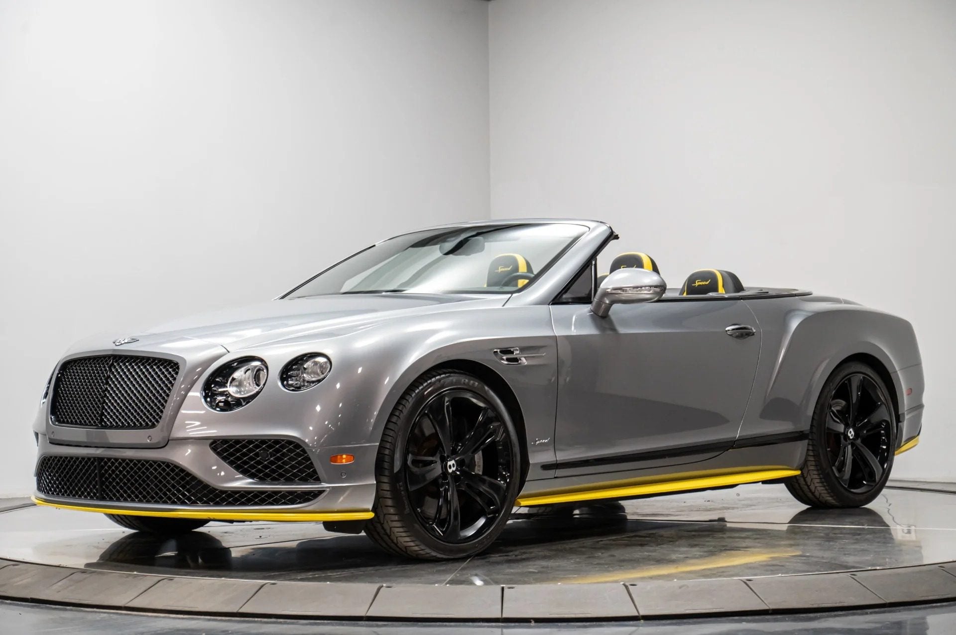 A silver Bentley Continental GTC convertible with black wheels and yellow accents is parked indoors on a rotating platform, embodying the essence of a refined luxury tourer.