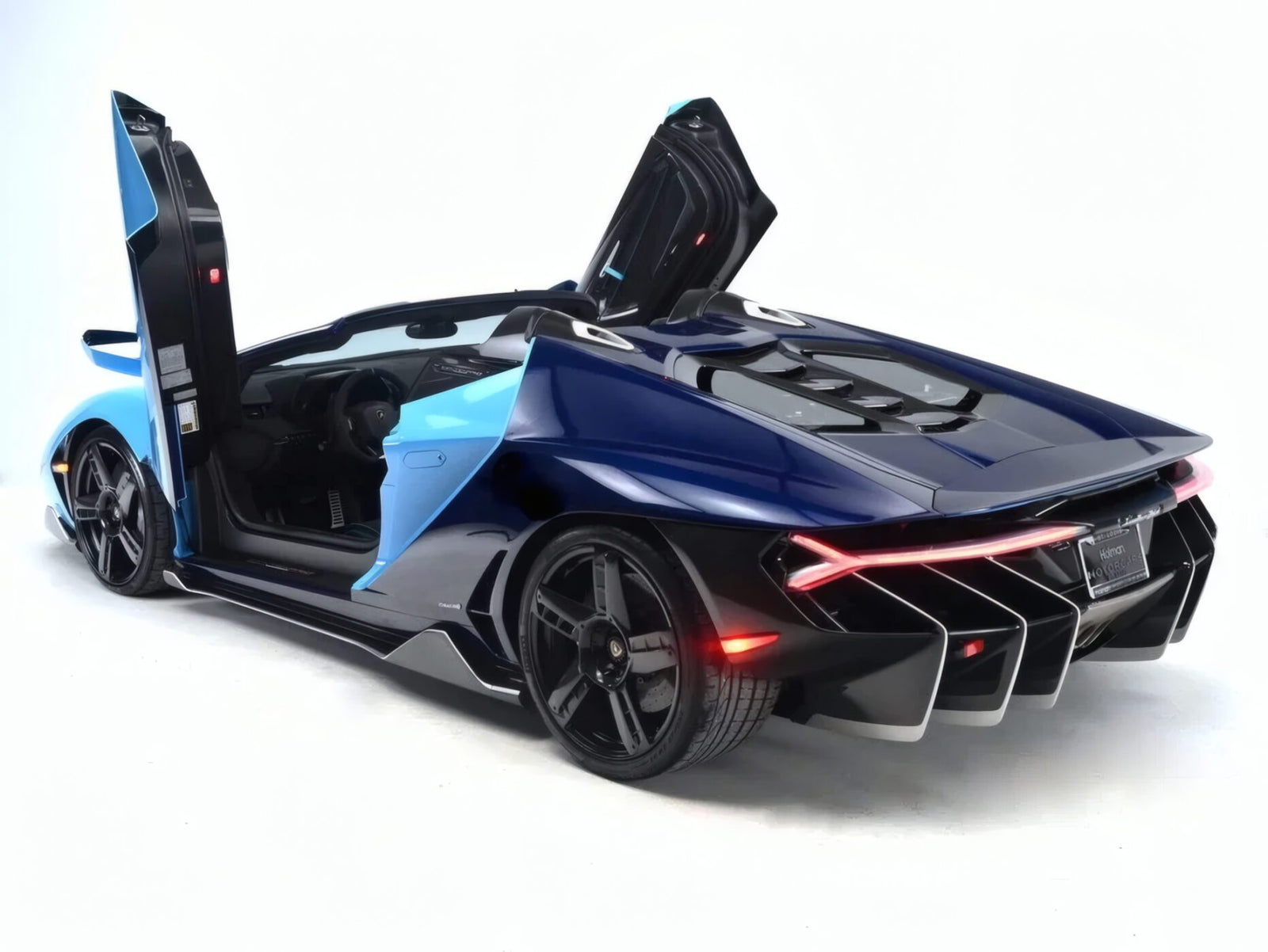 A blue and black Lamborghini Centenario Roadster with its scissor doors open, viewed from the rear on a white background. Only 10 in the U.S.: this luxury sports car is an ultra-rare masterpiece hitting the market.