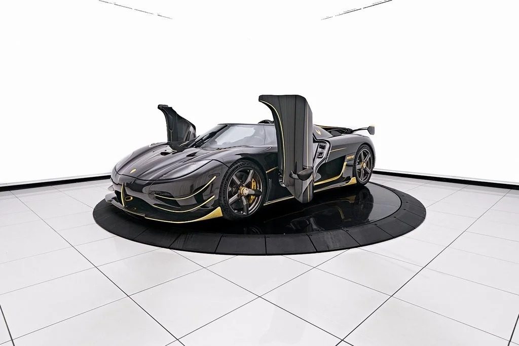 A sleek, black 2018 Koenigsegg with gull-wing doors open, displayed on a circular platform in a well-lit showroom with a white background.
