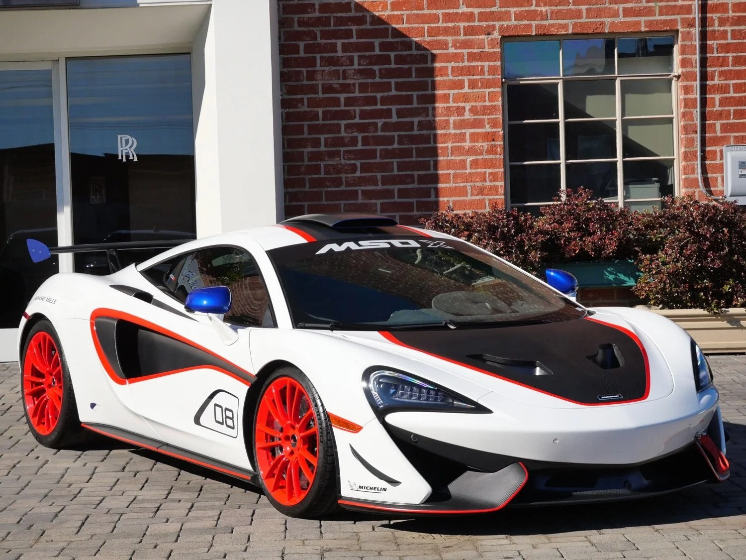 A white supercar with red accents and wheels is parked on a cobblestone driveway in front of a brick building and a glass door, hinting at its undeniable racing pedigree.