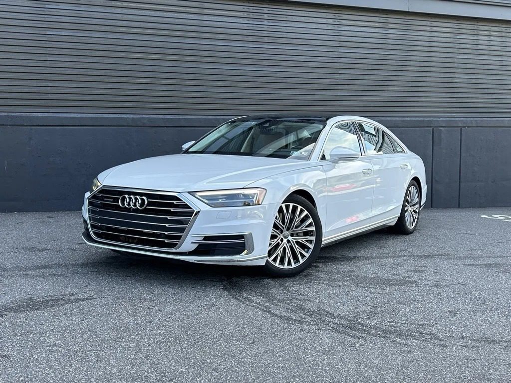 A white Audi sedan, a standout among affordable luxury sedans, is parked on a paved surface in front of a dark, ribbed metal wall.