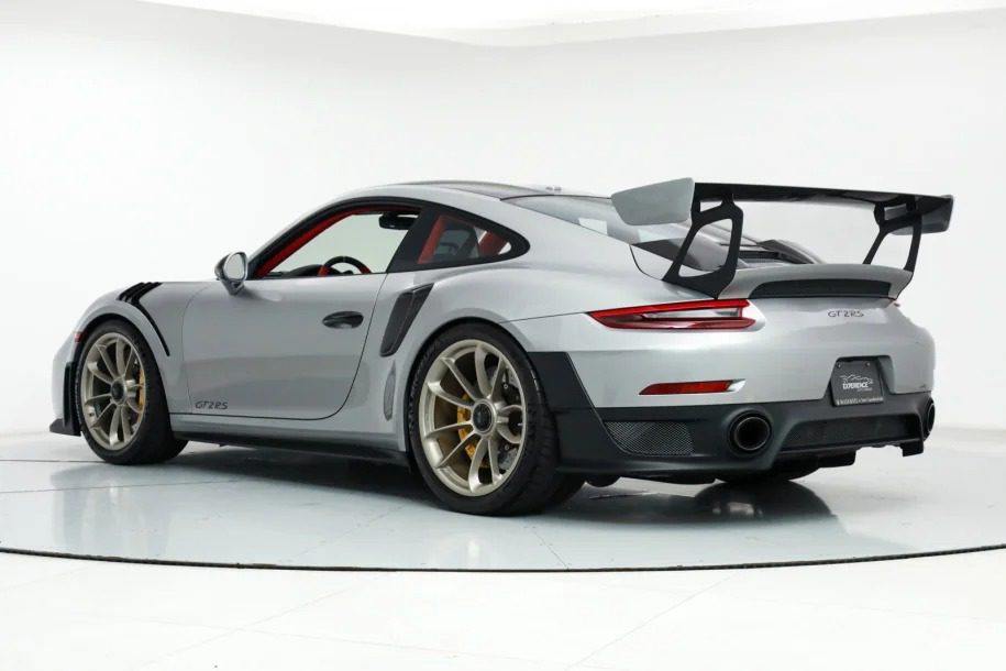 A silver Porsche GT3 RS sports car, known as the ultimate speed machine, features a large rear spoiler and gold alloy wheels, viewed from the rear three-quarter angle in a white studio setting.