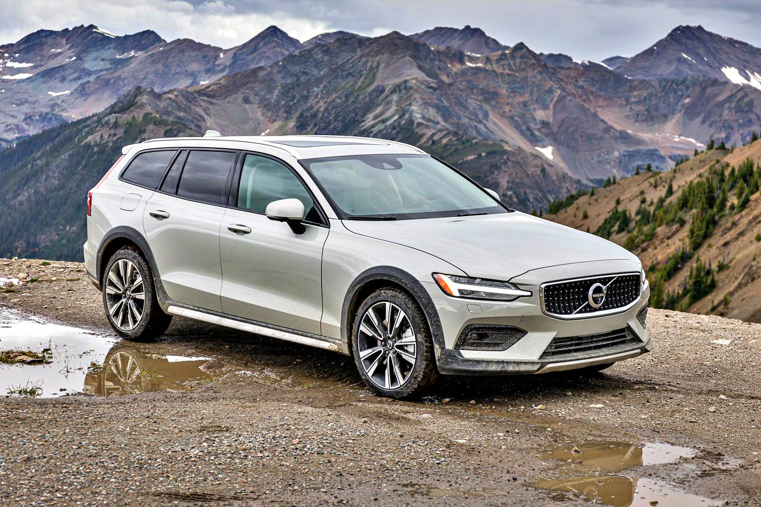 A silver Volvo V60 Cross Country wagon is parked on a gravel area near a mountain range on a cloudy day.