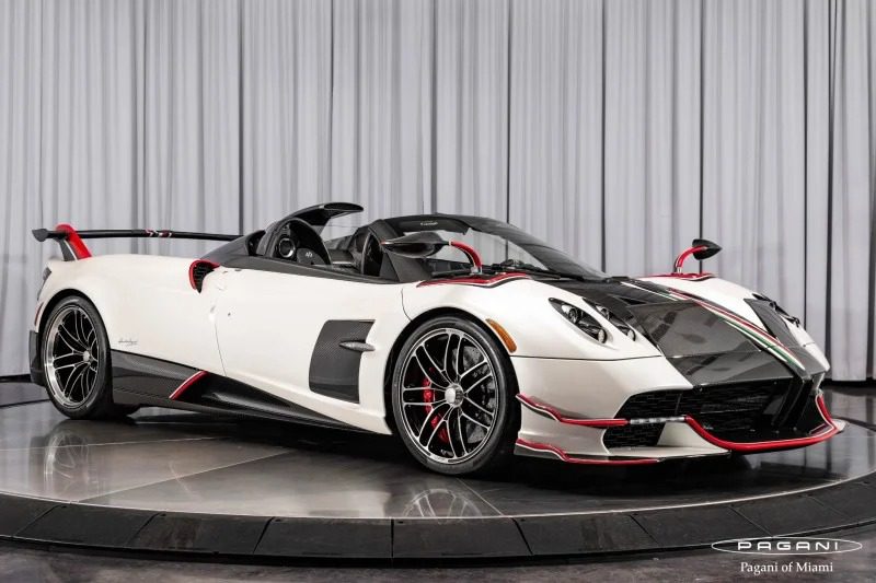 A white and black Pagani Huayra with red accents, featuring an aerodynamic design and convertible roof, is showcased on a rotating platform in the showroom. This stunning Roadster BC is for sale to those who crave elegance and performance.