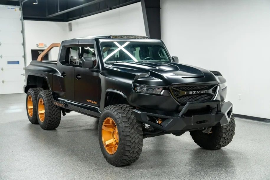 The black Rezvani Hercules 6x6 truck, featuring large off-road tires and gold rims, is parked indoors with a rugged design and a visible roll bar at the back. This impressive vehicle is now for sale.