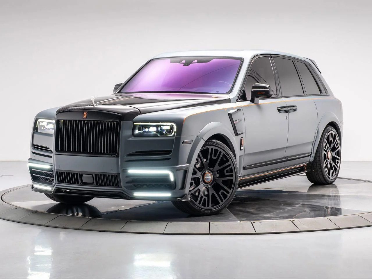 The Rolls-Royce Cullinan, a luxury SUV with a matte silver and black finish, large black wheels, tinted windows, and LED headlights, is elegantly displayed in a studio setting on a rotating platform.