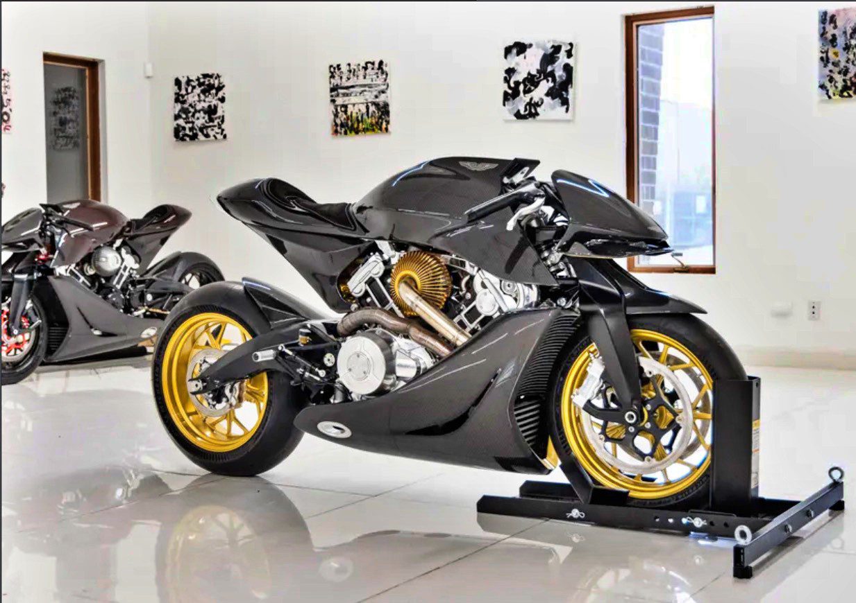 A sleek black Aston Martin AMB 001 motorcycle with gold wheels is displayed on a stand inside a modern showroom with white walls, windows, and art. Another motorcycle is in the background at Sotheby’s Motorsport in 2022.