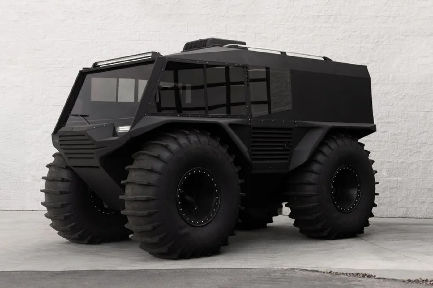 A futuristic black all-terrain vehicle with large tires is parked on a concrete surface against a white brick wall.