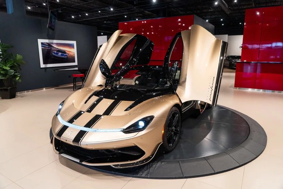 A gold and black electric hypercar with its scissor doors open is displayed in a showroom with a red and black backdrop, highlighting the stunning Pininfarina Battista for sale.