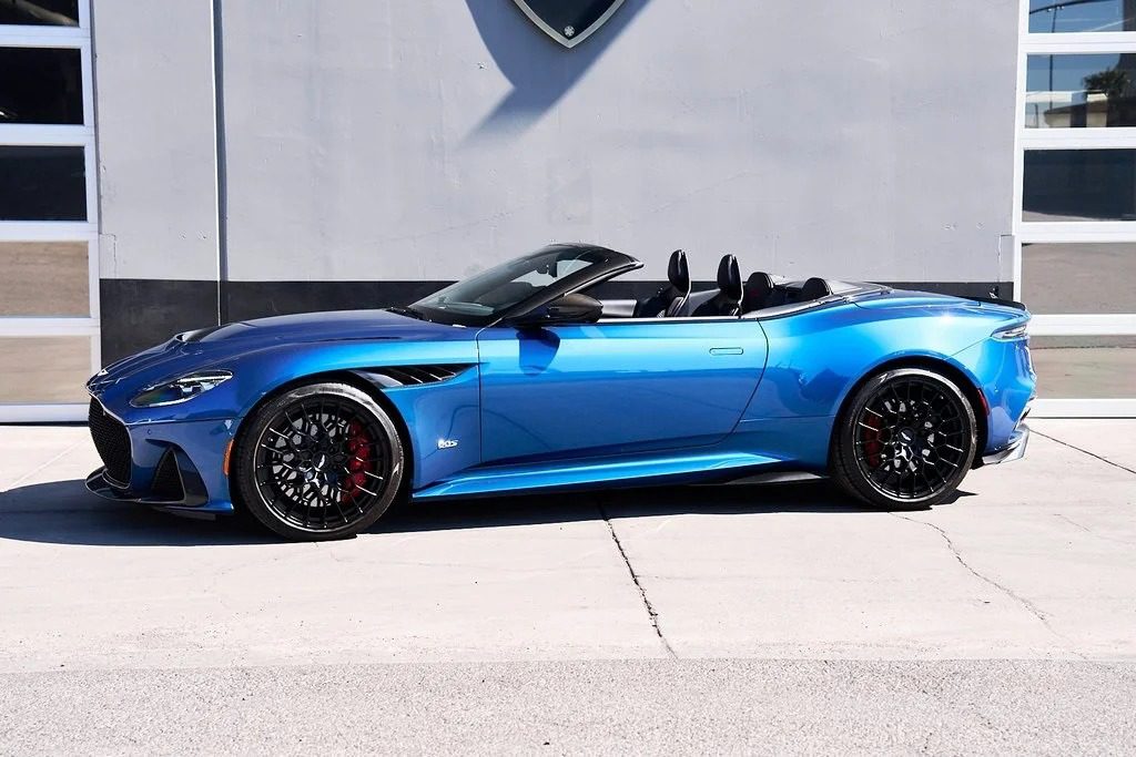 A blue Aston Martin DBS 770 Ultimate Grand Touring masterpiece is parked in front of a gray building with large windows and a garage door.