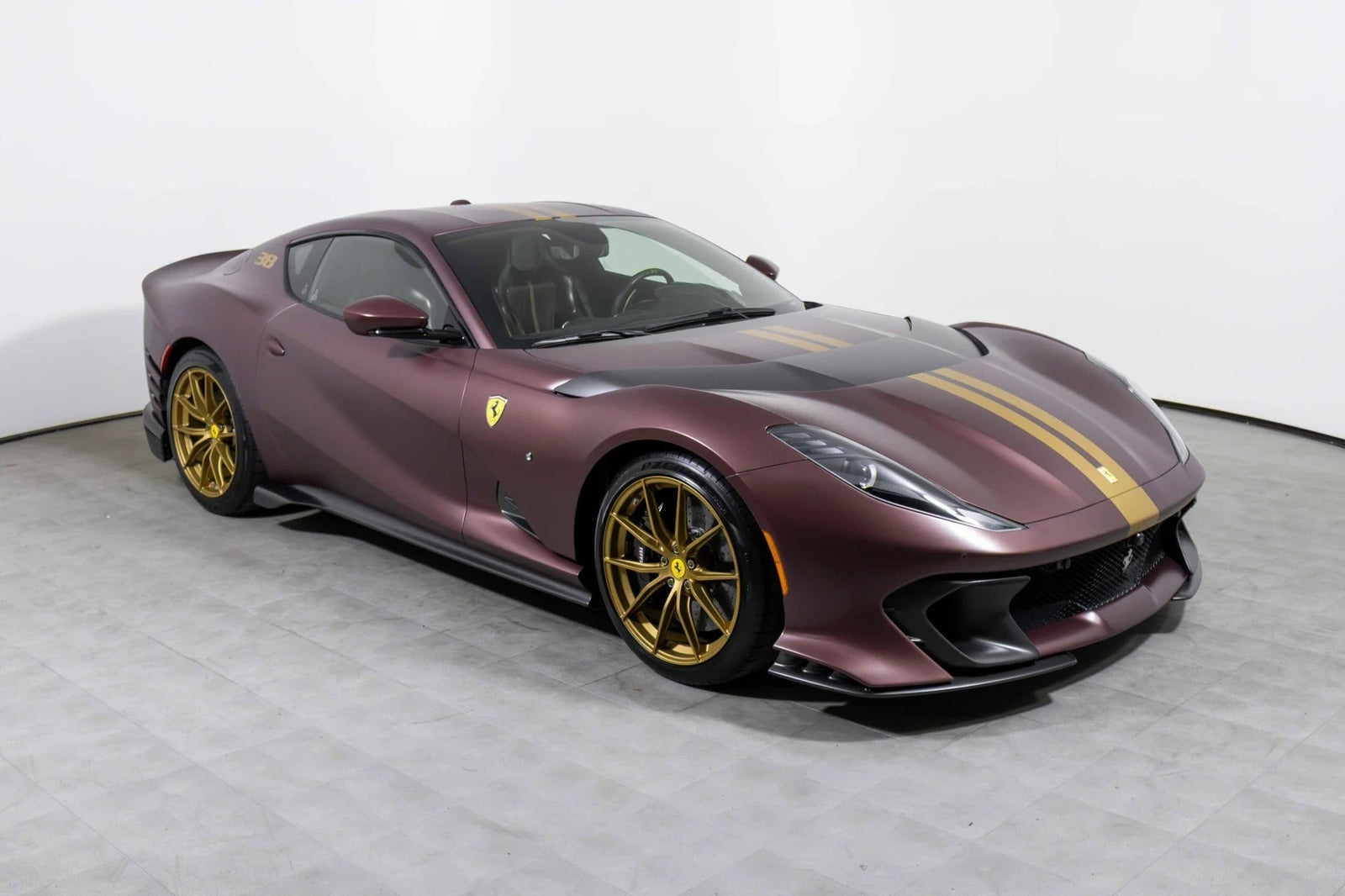 A maroon Ferrari 812 Competizione with gold racing stripes and V12 power, featuring gold wheels, is parked indoors on a gray tiled floor against a white wall.