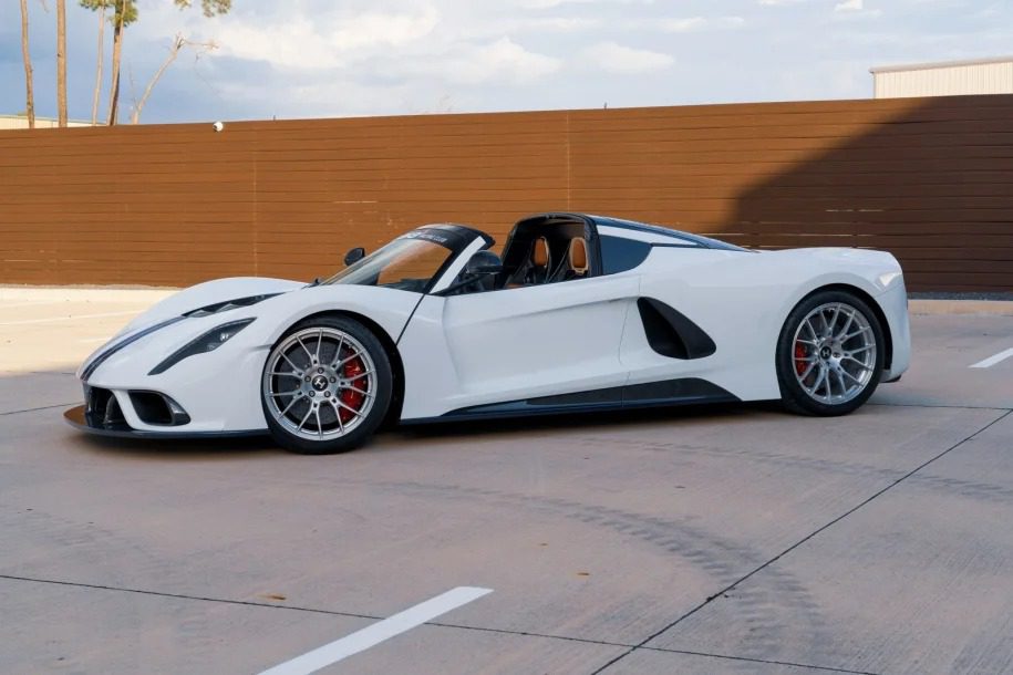 A sleek white roadster is parked on a concrete surface, with a rustic wooden fence in the background, exuding the allure of a Heritage Series masterpiece.