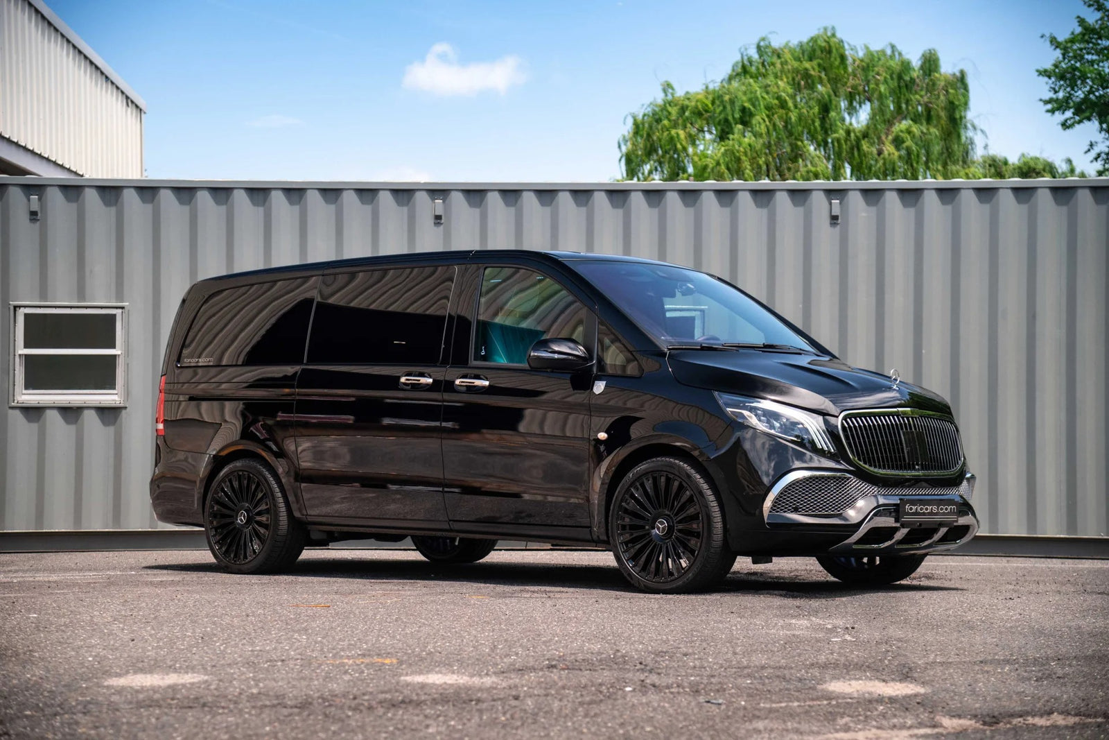 A black luxury van with tinted windows and black alloy wheels is parked on pavement in front of a gray shipping container, showcasing the premium appeal of FariCars' luxury vans.