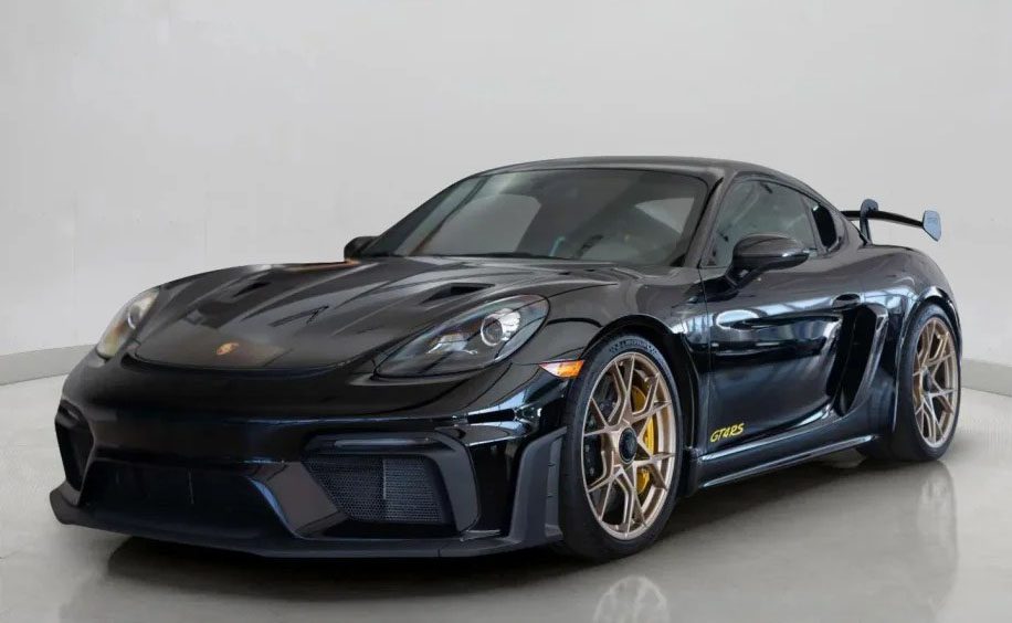 A sleek black Porsche 718 Cayman GT4 RS with gold rims and a rear spoiler is on display in the showroom. Track-ready and stunning, this sports car is now for sale.