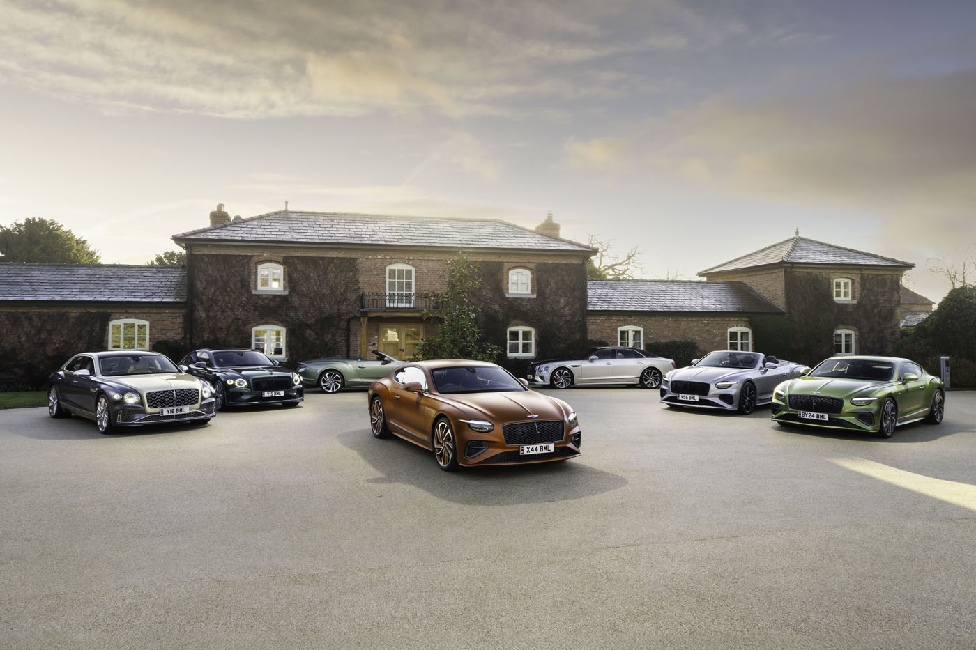 A group of luxury 2024 Bentleys is parked in front of a large stone house on a cloudy day.